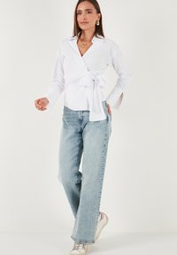 Woman wearing a white long-sleeve wrap blouse with side bow, light blue straight-leg jeans, and white sneakers against a plain background.