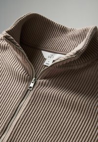 Giacca zip-up marrone a coste con colletto alto e cerniera argento. Include un'etichetta con il marchio e la taglia.