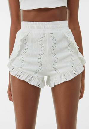 Hvide bomuldsshorts med elastisk taljebånd, øjenslyngemønstre og flæsekant, båret af en person fra taljen til midt på låret.