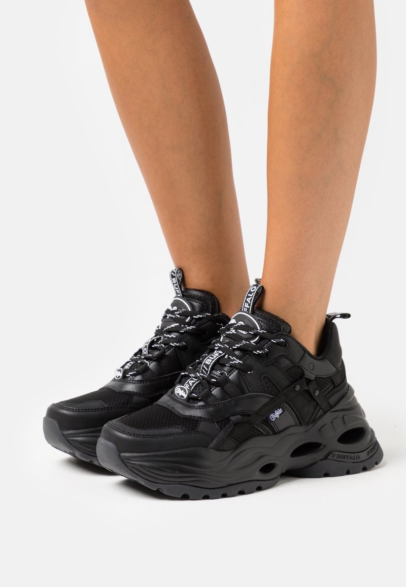 Baskets noires épaisses avec semelles épaisses, détails en mesh et cuir, lacets blancs à motifs, portées pieds nus sur un fond blanc.