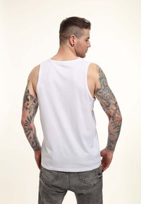 Hvid bomulds tanktop med rund halsudskæring og brede armhuller. Viser synlige tatoveringer på venstre arm, sat mod en neutral baggrund.