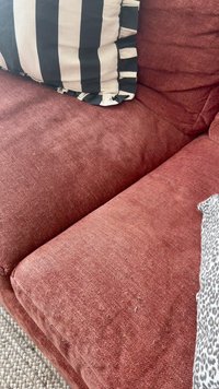 Sarkans auduma dīvāns ar teksturētu apdari, ar melnbaltu svītrainu spilvenu un pelēku teksturētu pledu spilvenu.