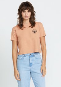 Krótkie, wycięte w talii t-shirt w kolorze brzoskwiniowym, wykonany z miękkiej bawełny, z krótkimi rękawami i czarną grafiką słońca z napisem "Volcom Stone". Połączony z jasnoniebieskimi dżinsami.