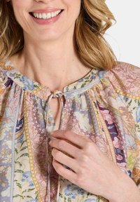 Blumenbluse mit rundem Ausschnitt und Bindeschluss. Aus pastellfarbenem Stoff, mit gerafften Ärmeln und komplizierten Mustern im gesamten Stoff.