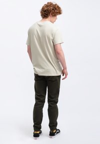 Lichtbeige katoenen T-shirt met een ronde hals, weergegeven in de achteraanzicht. Gecombineerd met donkergroene, taps toelopende pantalons en zwarte sneakers met witte zolen.