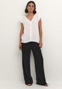 Blouse blanche à manches courtes et col en V avec des points texturés et des boutons sur le devant, associée à un pantalon noir à jambes larges en matière fluide.