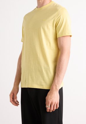 Mand iført gul t-shirt med korte ærmer og sorte bukser, står med armene afslappet langs siden mod en ensfarvet baggrund.
