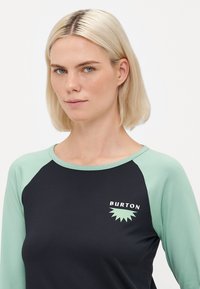 Blonda sieviete ar zilām acīm, ģērbusies melnā un piparmētru zaļā garajās piedurknēs krekli, ar "BURTON" logo uz krūtīm.