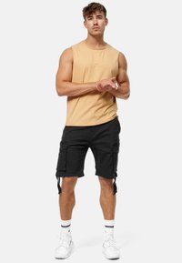 Chemise beige sans manches assortie à un short cargo noir. Le short présente des poches latérales et des sangles ajustables. Baskets blanches avec des rayures noires.
