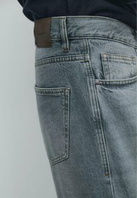 Jasnoniebieskie dżinsy z denimu o lekko spłowiałej fakturze, wyposażone w standardowe kieszenie oraz brązową skórzaną naszywkę na pasie.