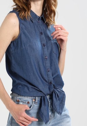 Ærmeløs blå denim skjorte med knapper, bundet i taljen, båret med lyseblå jeans på en person med bølget lyst brunt hår.