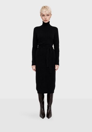 Femme aux cheveux blonds courts portant une robe noire à manches longues avec un col roulé, une ceinture à la taille et des bottes sombres pointues montantes jusqu'aux genoux, debout devant un fond uni.