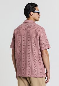 Rosa Kurzarmhemd mit Diamantmuster und offenem Strickdesign, ausgestattet mit einem klassischen Kragen und einem entspannten Schnitt. Detaillierte Textur sichtbar.