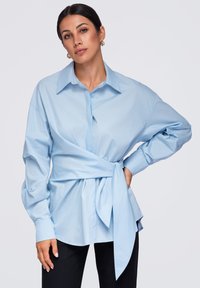 Femme portant une chemise bleu clair à manches longues avec une grande ceinture à la taille, associée à un pantalon noir, posant avec une main sur la hanche.