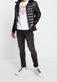 Hombre con chaqueta de puffer negra, sudadera blanca de Guess con logo rojo, jeans negros y zapatillas blancas con cordones negros, de pie sobre un suelo blanco.