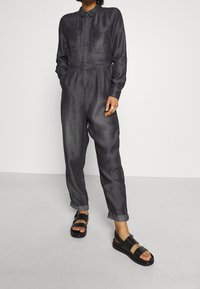 Mono gris oscuro con cuello, dos bolsillos delanteros, cintura elástica, piernas entalladas, combinado con sandalias negras de tiras gruesas.