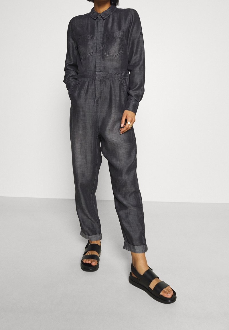 Mono gris oscuro con cuello, dos bolsillos delanteros, cintura elástica, piernas entalladas, combinado con sandalias negras de tiras gruesas.