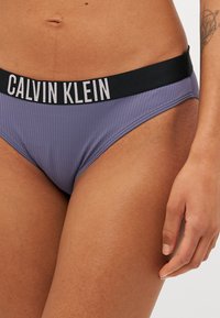 Bikini Calvin Klein iz rebraste vijolične tkanine in črnimi elastičnimi pasovi s belim napisom. Brezšivni robovi povečujejo udobje.