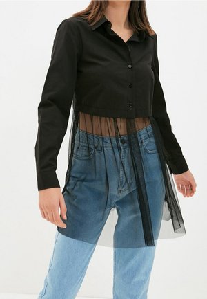 Vrouw die een zwart overhemd met lange mouwen en knopen draagt, met een doorschijnend mesh dat onder de taille doorloopt, gecombineerd met een hoog uitgesneden blauwe spijkerbroek op een witte achtergrond.
