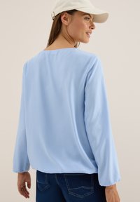 Vrouw in een lichtblauwe blouse met lange mouwen, donkere spijkerbroek en een witte baseballpet, die over haar schouder kijkt.