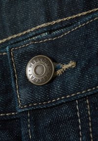 Tamnoplava traper tkanina s metalnim gumbom urezanim s natpisom "WEEKDAY", ojačanim šavovima i teksturiranom površinom.