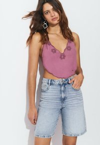 Top corto morado con bordado floral y dobladillo de forma ondulada, combinado con pantalones cortos de mezclilla azul claro. Modelo llevando pendientes geométricos.