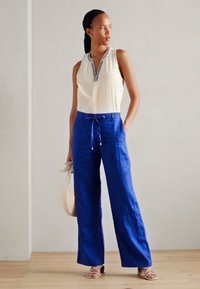 Woman wearing sleeveless white top and high-waist blue wide-leg pants, holding a beige bag, standing on wooden floor.