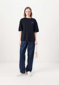 Navy blue short-sleeve t-shirt with relaxed fit and embroidered logo, paired with wide-leg denim jeans and white sneakers, holding a pink bag.