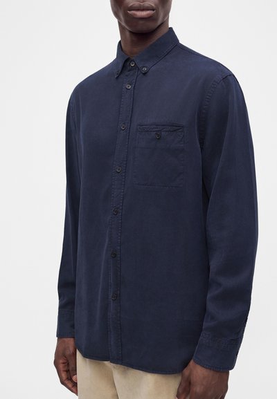 Man wearing a long-sleeve, dark blue button-up shirt with a chest pocket, standing against a plain light background.