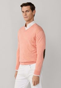 Mężczyzna ubrany w różowy sweter z dekoltem w serek nałożony na białą koszulę z kołnierzykiem i białe spodnie, stojący na tle jednolitego jasnoszarego tła.