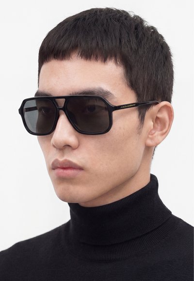 Jeune homme portant de grandes lunettes de soleil noires rectangulaires et un pull à col roulé noir sur un fond clair uni.