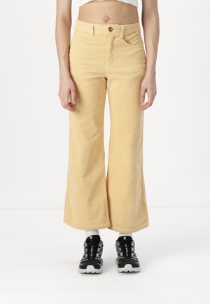Person trägt beige, hoch taillierte Cord-Hose mit weitem Bein, kombiniert mit schwarz-weißen Sportschuhen und weißen Socken.