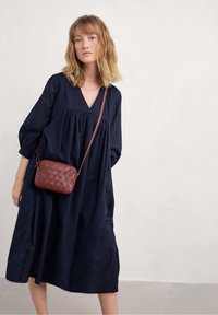 Tamsiai mėlyna suknelė su V formos iškirpte ir pūstomis rankovėmis, derinama su mažu ruduo pynimo odiniu krepšiu per petį. Lygus audinio tekstūra.