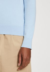 Maglione a maglia blu chiaro con orlo e polsini a coste, che mette in mostra una superficie liscia. La mano poggia su un tessuto beige.