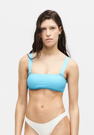 Mladá žena s mokrými vlasmi v modrom športovom športovom podprsenke a bielych spodných dieloch bikín na jednoduchom svetlom pozadí.