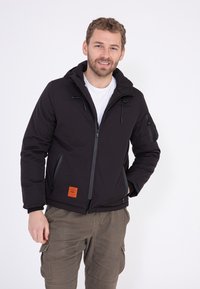 Zwarte geïsoleerde jas met capuchon, frontale rits, zijzakken en een oranje label patch. Beschikt over een gladde, matte textuur en verstelbare trekkoorden.