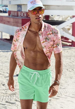 Kurzärmliges, pinkfarbenes Hemd mit Palmblättern, vorne offen. Hellgrüne Badeshorts mit weißem Kordelzug. Helle Sonnenbrille und Mütze mit Text.
