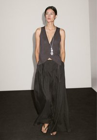 Dark, sleeveless top with a deep V-neck, paired with loose black pinstriped trousers. Long necklace with multiple oval pendants.