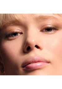 Inquadratura ravvicinata di un volto con capelli biondo chiaro, tocchi di luce sulla pelle e labbra rosa lucide. L'ombretto in tonalità neutre esalta lo sguardo.