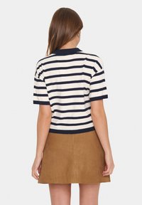 Donna con lunghi capelli castani che indossa una maglietta a righe bianche e navy a maniche corte e una gonna in suede color sabbia, vista di schiena.