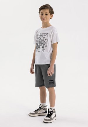 Junge steht, trägt weißes T-Shirt mit Grafik, dunkelgraue Shorts mit Patch, weiße Socken und graue High-Top-Sneaker vor weißem Hintergrund.