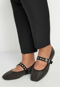 Bailarinas de ante negro con punta redonda, adornadas con tachuelas plateadas y una correa en el tobillo con herrajes plateados. Combinadas con pantalones cortos negros.