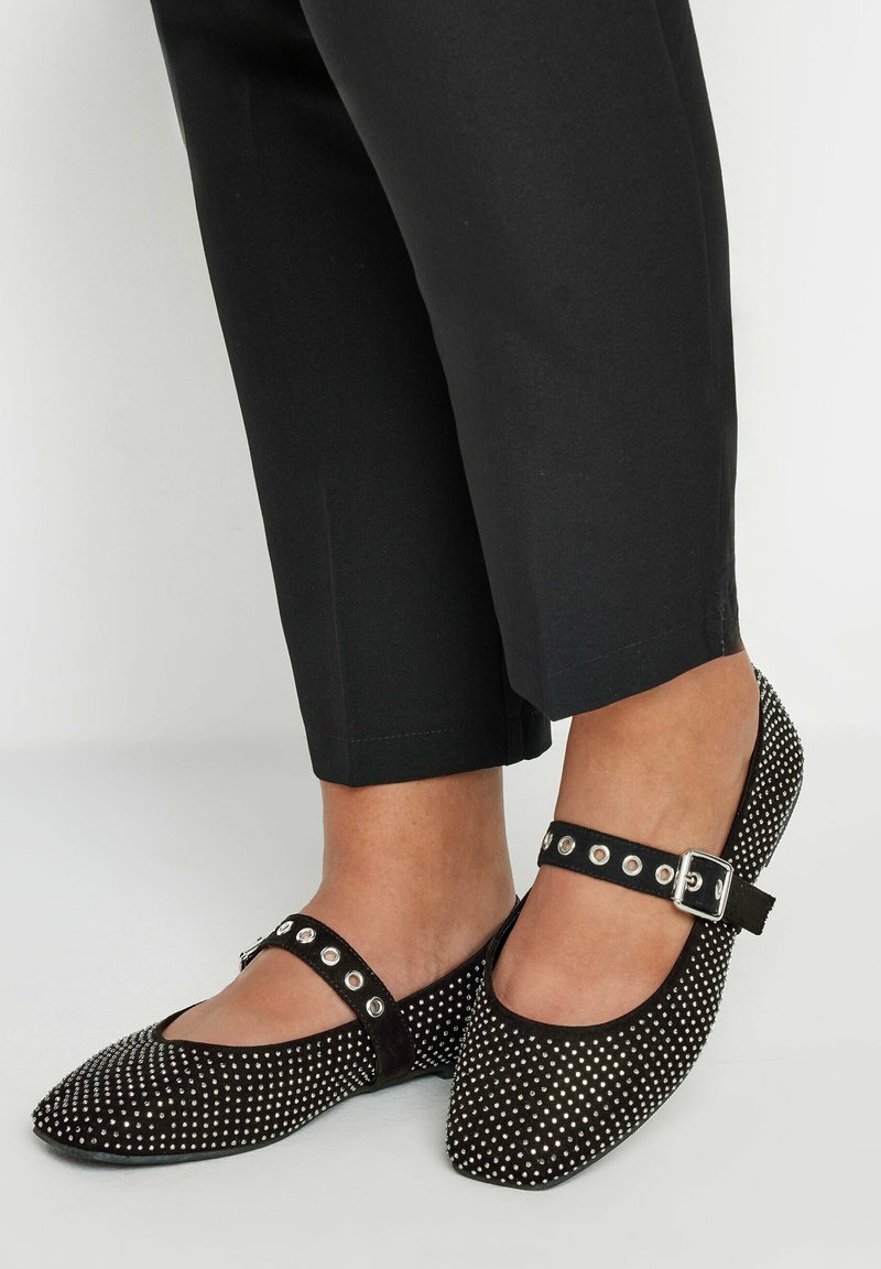 Bailarinas de ante negro con punta redonda, adornadas con tachuelas plateadas y una correa en el tobillo con herrajes plateados. Combinadas con pantalones cortos negros.