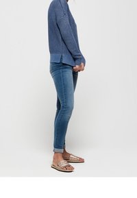 Blue knitted sweater with textured pattern, side slits at hem. Paired with light blue skinny jeans and beige sandals with woven accents.