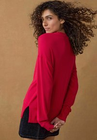 Maglione rosso a maniche lunghe con orlo a coste e fessure laterali, caratterizzato da una vestibilità rilassata e un knit testurizzato. Indossato con una gonna scura.