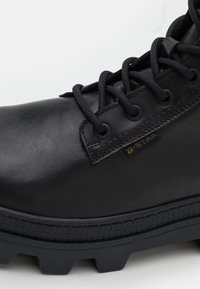 Botín de cuero negro con cordones redondos, ojetes metálicos y suela de goma texturizada con patrones de relieve prominentes. Logo de G-Star visible.