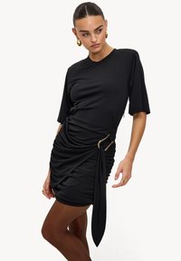 Robe noire à manches courtes, avec un tissu froncé, une silhouette drapée et un ornement en métal doré. Matière douce et extensible.