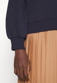 Pullover blu navy con maniche a sbuffo e polsini a costine. Abbinato a una gonna pieghettata color sabbia, che mostra tessuti lisci e testurizzati con un design fluido.