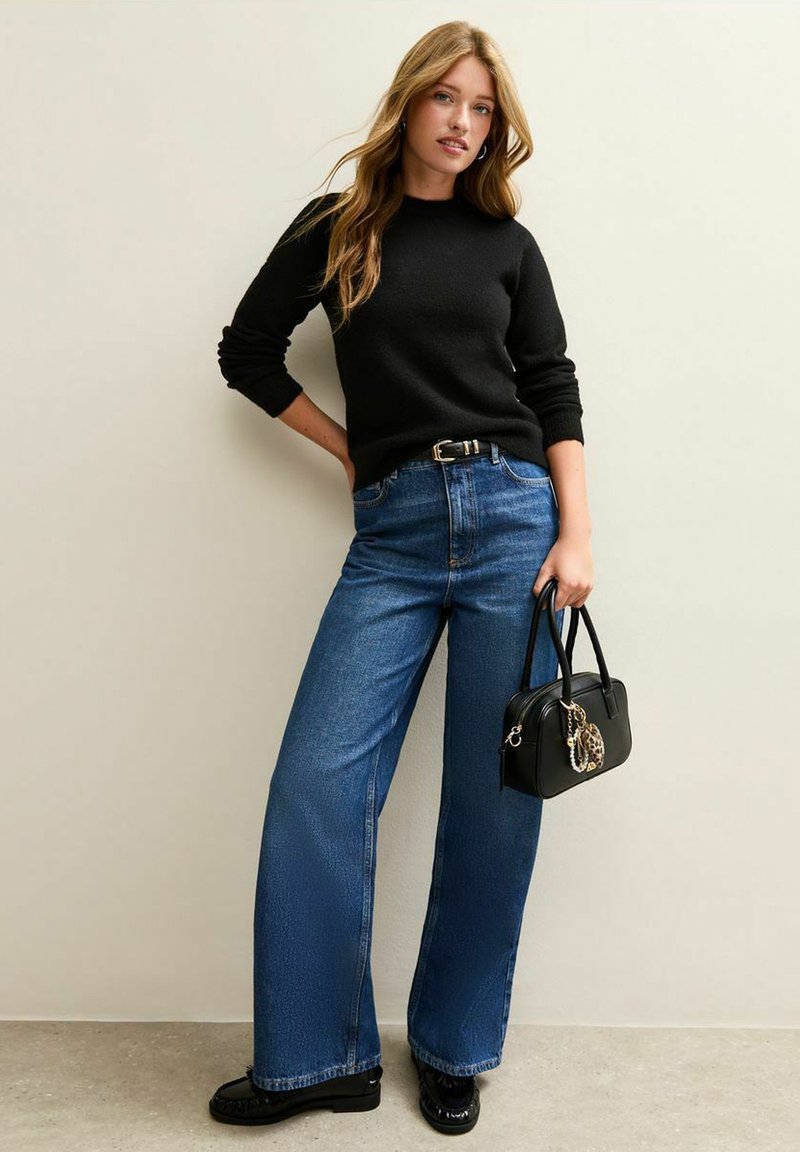 Schwarzer Pullover, hoch-taillierte blaue weite Jeans und eine kleine schwarze Handtasche mit Akzent. Das Outfit zeichnet sich durch minimalistisches Design und Texturen aus.