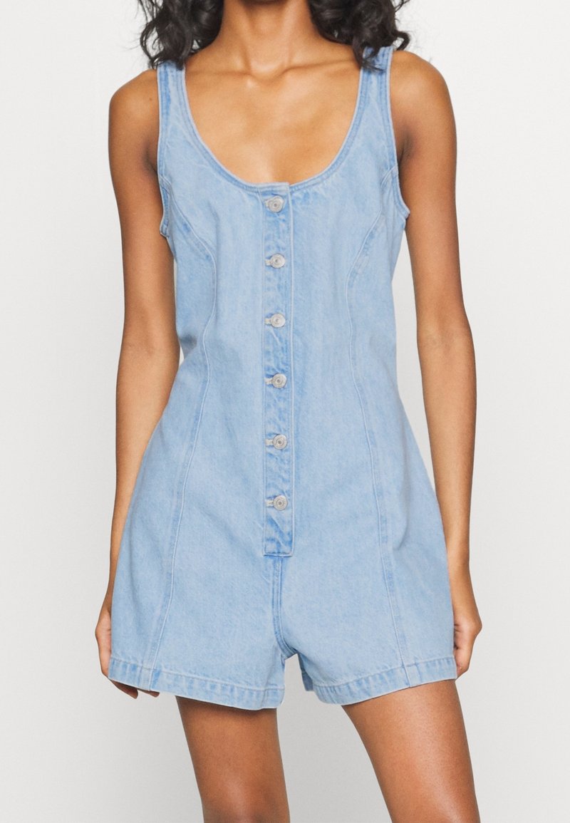 Femme portant une combinaison courte sans manches en denim bleu clair avec fermeture à boutons sur le devant et un short, debout devant un fond blanc uni.